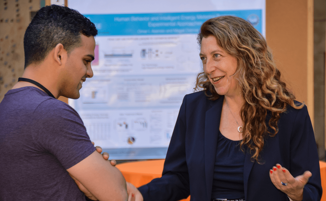 The photo shows two people talking (student and administrator) in front of a research poster.